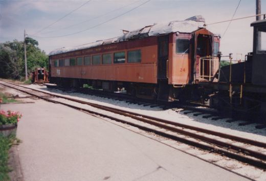 A vintage orange and maroon interurban railcar, numbered 24 and lettered for the Chicago South Shore and South Bend Railroad, is parked on tracks in a sunny yard with a damaged roof.