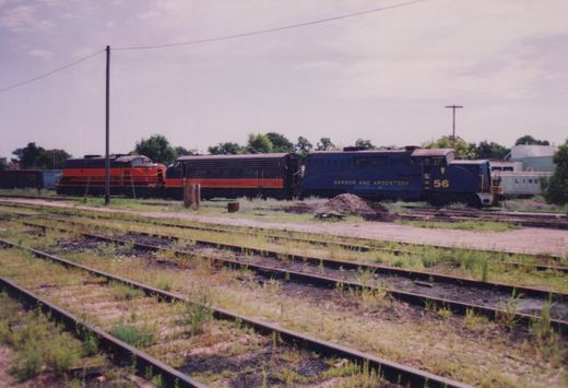 A blue Bangor and Aroostook locomotive #56 and an orange and black Wisconsin & Calumet locomotive #52 are parked on parallel tracks in a grassy railroad yard under an overcast sky.