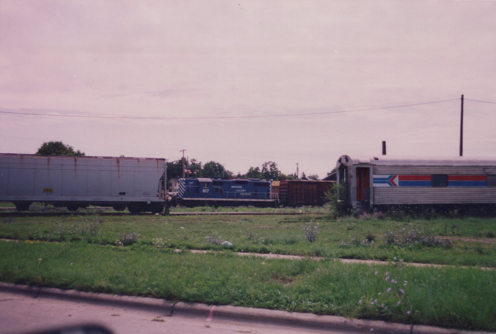 A blue Wisconsin Calumet Railroad locomotive numbered 617 is parked in a grassy yard next to an Amtrak passenger car and a gray boxcar under an overcast sky.