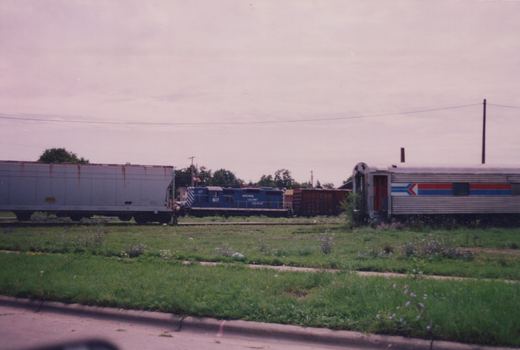 A blue Wisconsin Calumet Railroad locomotive numbered 617 is parked in a grassy yard next to an Amtrak passenger car and a gray boxcar under an overcast sky.
