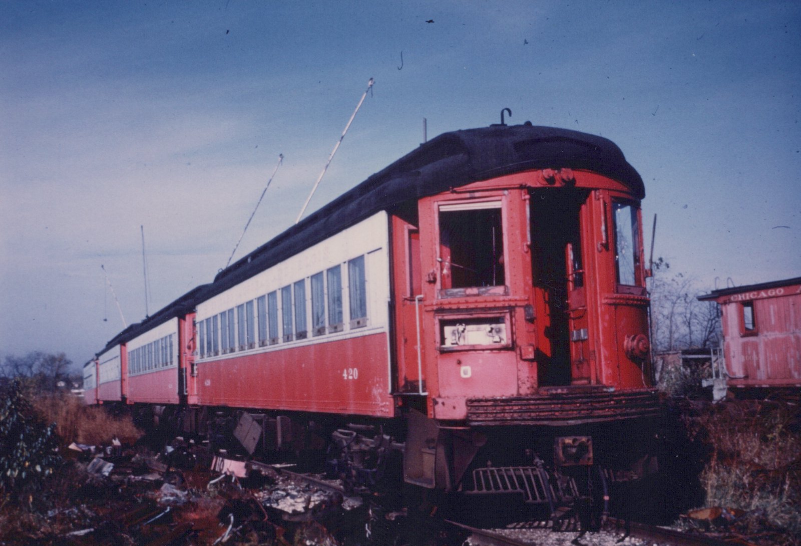 A red and cream interurban railcar, numbered 420, is shown from the rear three-quarter view, sitting on tracks with a decaying caboose labeled 'CHICAGO' in the background under an overcast sky.
