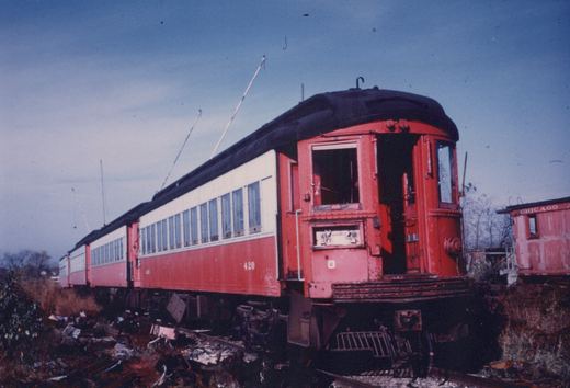 A red and cream interurban railcar, numbered 420, is shown from the rear three-quarter view, sitting on tracks with a decaying caboose labeled 'CHICAGO' in the background under an overcast sky.