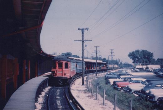 An electric interurban train with red front and cream sides, numbered 634, is on a curved track beside a wooden platform, with a parking lot full of 1950s cars to the right.