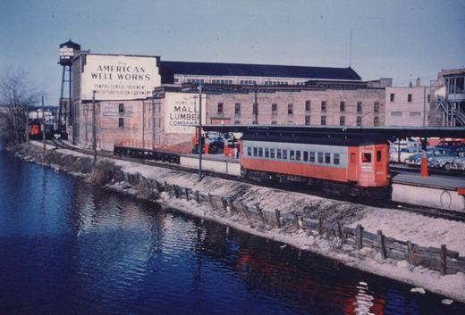 An orange and silver interurban train car labeled 'Chicago Aurora and Elgin' and numbered 414 is stopped at a station platform beside a river in winter, with industrial buildings and parked cars visible in the background.