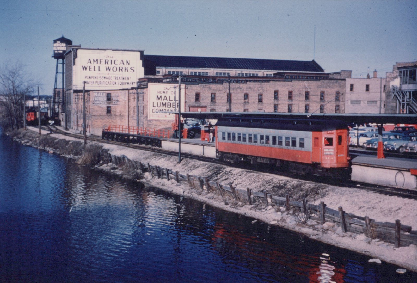 An orange and silver interurban train car labeled 'Chicago Aurora and Elgin' and numbered 414 is stopped at a station platform beside a river in winter, with industrial buildings and parked cars visible in the background.