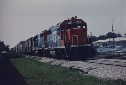 A red, gray, and white Canadian National locomotive, number 6418, pulling a long line of freight cars along a gravel track next to a grassy area and a parking lot with a fence.