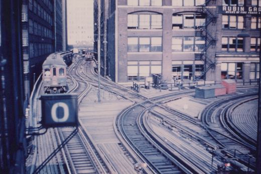 A vintage elevated train with a cream and maroon livery travels on wooden-decked tracks between tall buildings, passing a signal marked '0' and a building with a 'Rubin Grand' sign.