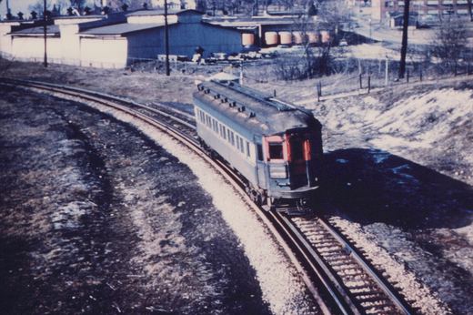 A blue and white streamlined interurban car, number 716, travels on a curved track through a snowy, industrial landscape.
