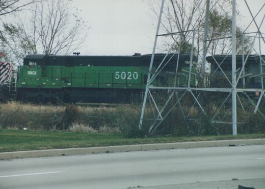 A green diesel locomotive numbered 5020 with RMGX lettering is on a track beside a road, with bare trees and an overcast sky in the background.