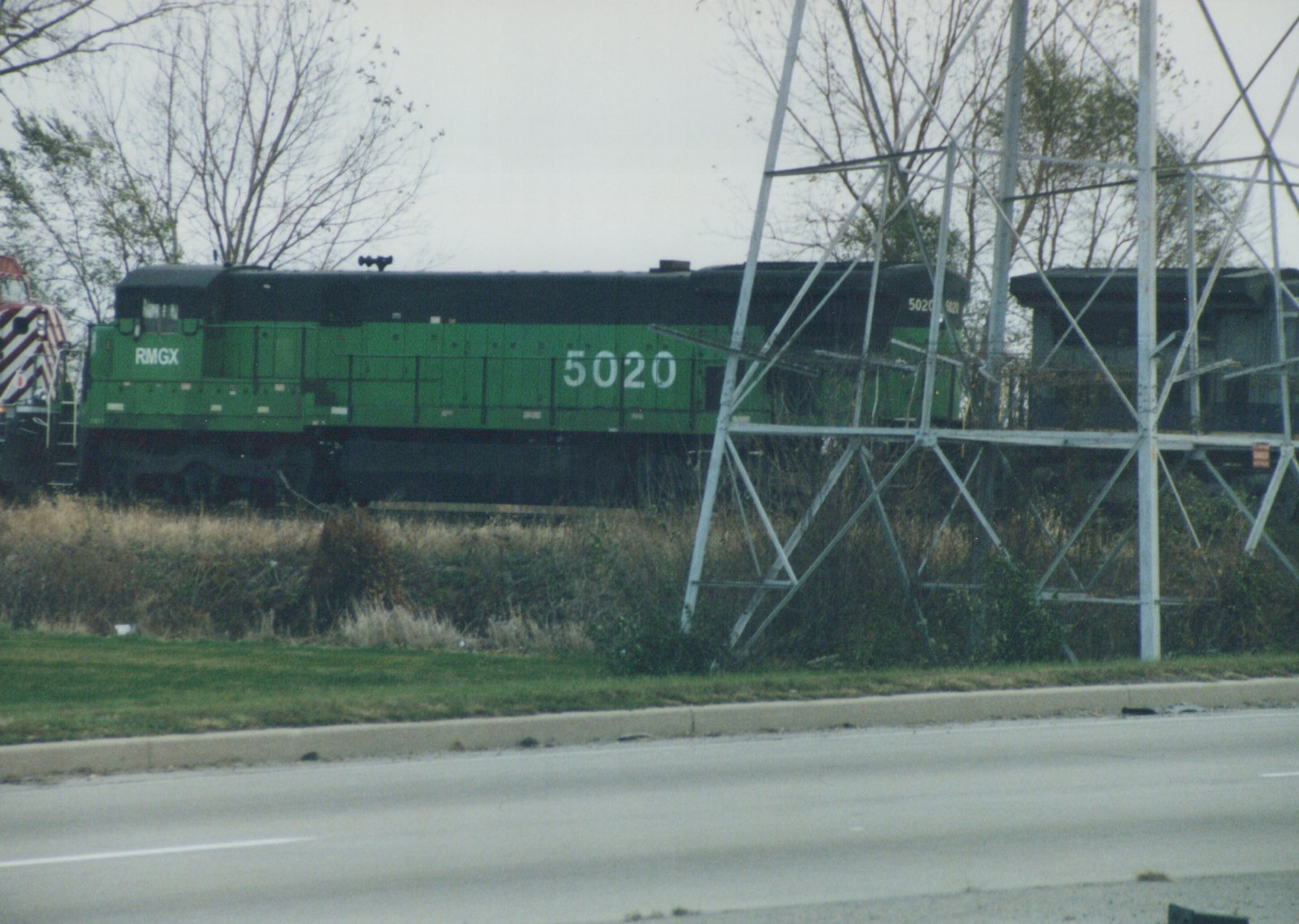 A green diesel locomotive numbered 5020 with RMGX lettering is on a track beside a road, with bare trees and an overcast sky in the background.