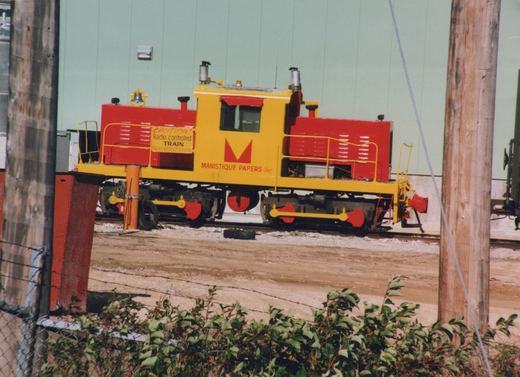 A yellow and red diesel locomotive with 'Manistique Papers Inc' lettering sits on tracks beside a light green building, viewed from a fenced area with greenery in the foreground.