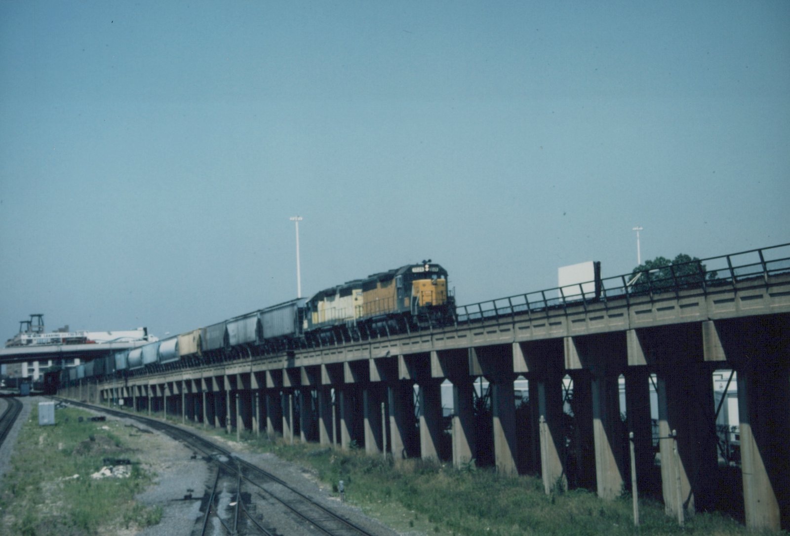 A long freight train on a concrete elevated bridge under a clear blue sky, with railroad tracks and grassy embankment below.