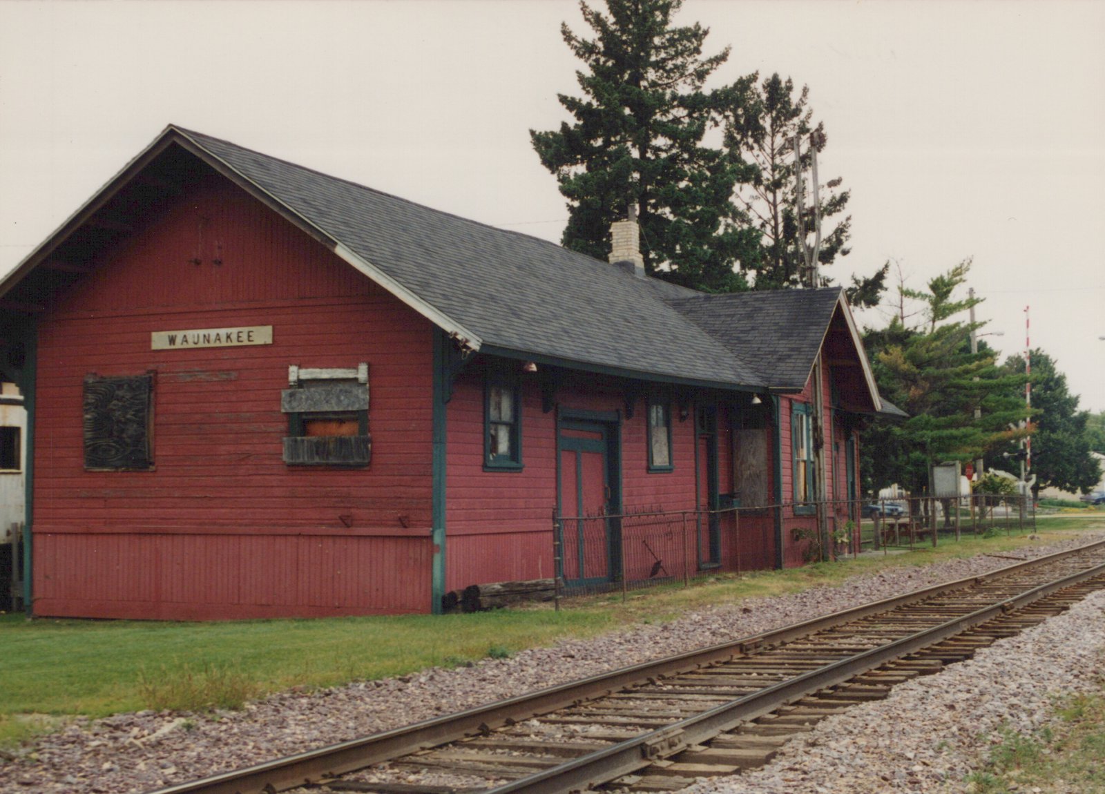 A red wooden railroad depot with a sign reading 'WAUNAKEE' sits beside a single set of tracks, with some windows boarded up and a blue car parked in the distance.
