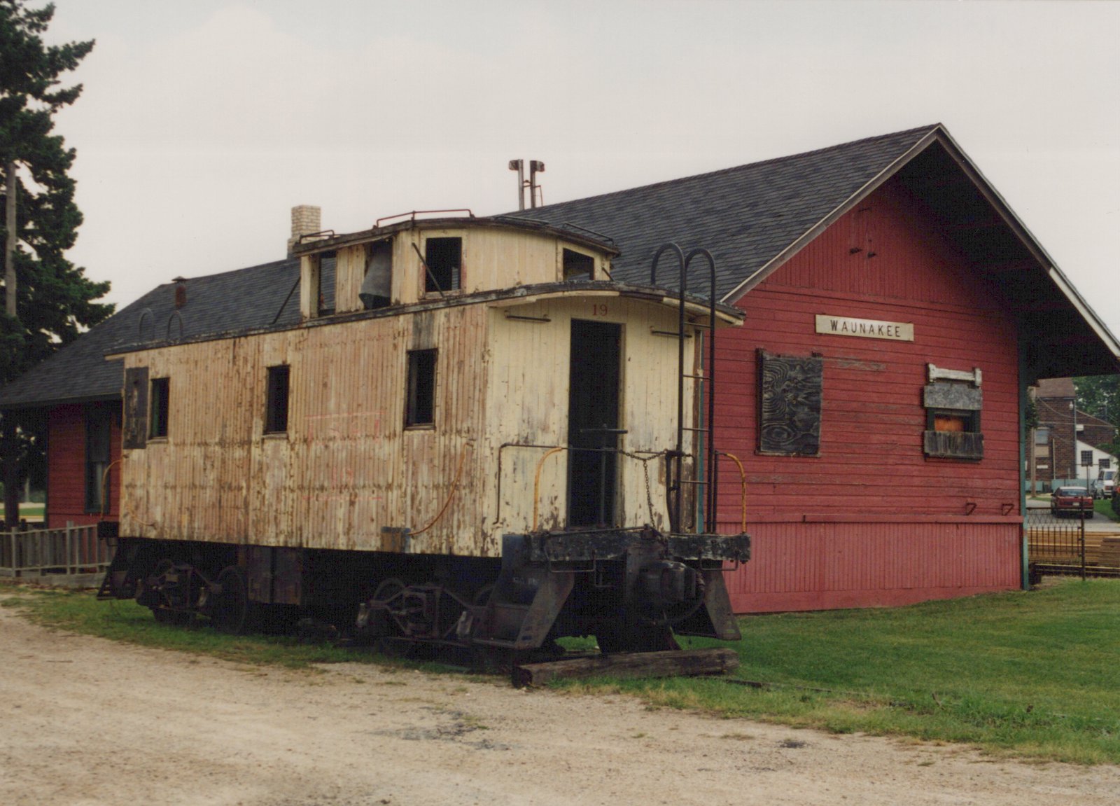 An old, cream-colored wooden caboose with the number 19 on its side is parked next to a red station building labeled 'WAUNAKEE', with a gravel foreground and an overcast sky.