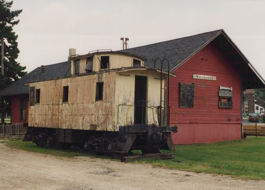 An old, cream-colored wooden caboose with the number 19 on its side is parked next to a red station building labeled 'WAUNAKEE', with a gravel foreground and an overcast sky.