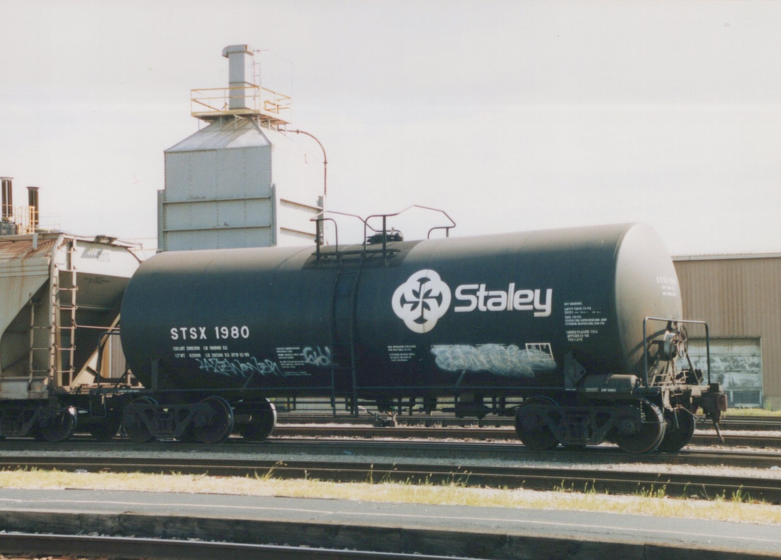 A side view of a dark green Staley tank car with the reporting mark STSX 1980, parked on tracks in an industrial yard under an overcast sky.