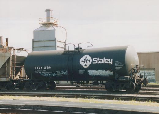 A side view of a dark green Staley tank car with the reporting mark STSX 1980, parked on tracks in an industrial yard under an overcast sky.