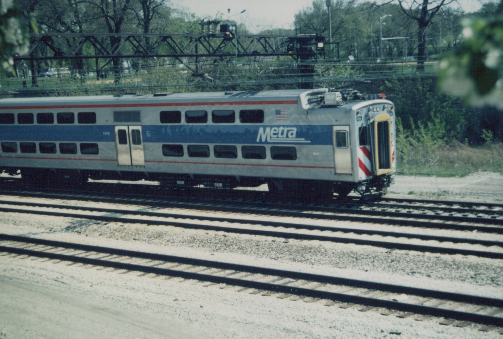 A silver and blue electric commuter rail car, number 1519, branded with the Metra logo, sits on a set of tracks with overhead wires, surrounded by trees and infrastructure.