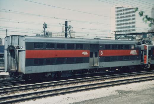 A silver and red Metra commuter train car, number 1536, sits on tracks with overhead wires and buildings in the background.