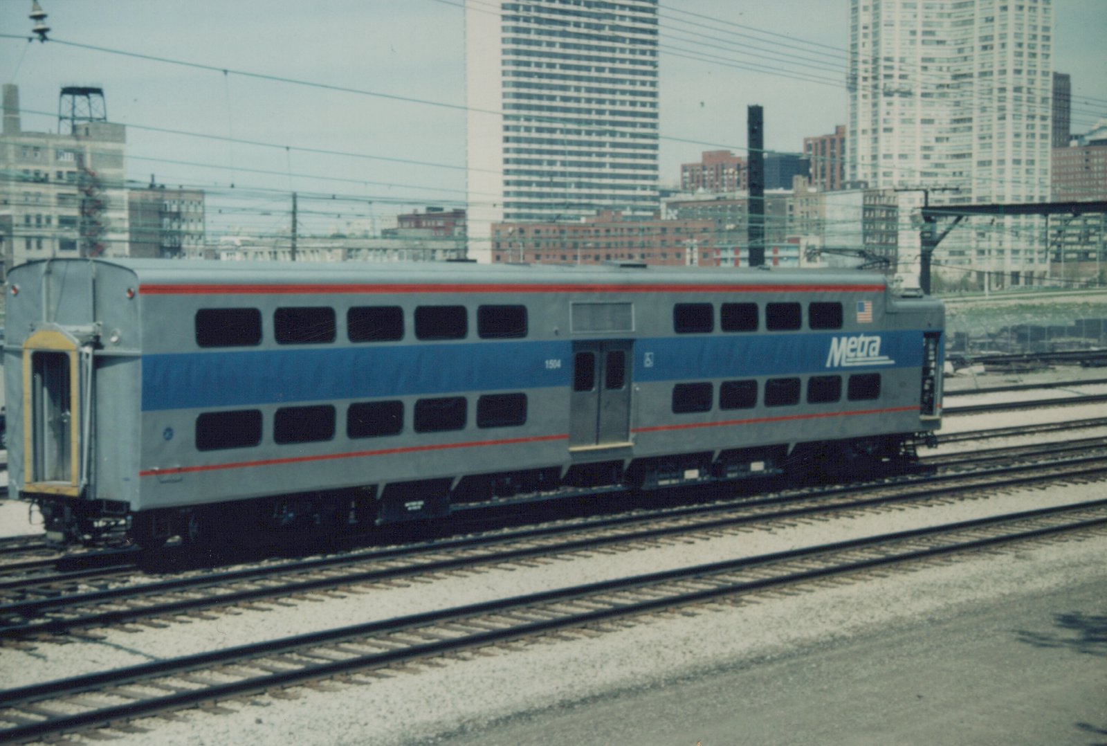 A gray and blue Metra Gallery commuter car numbered 1504 sits on railroad tracks with a city skyline behind it.
