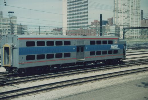 A gray and blue Metra Gallery commuter car numbered 1504 sits on railroad tracks with a city skyline behind it.