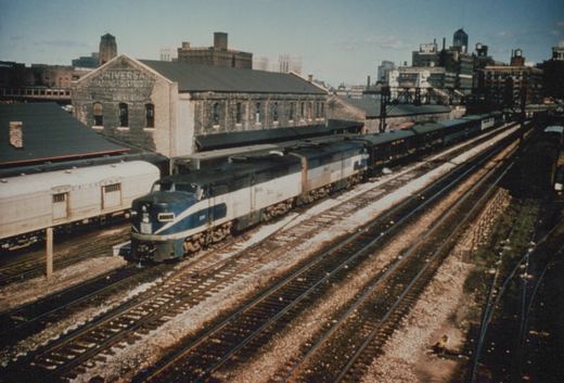 A blue and white diesel locomotive pulls a passenger train on multiple tracks, with a brick building and city skyline in the background.