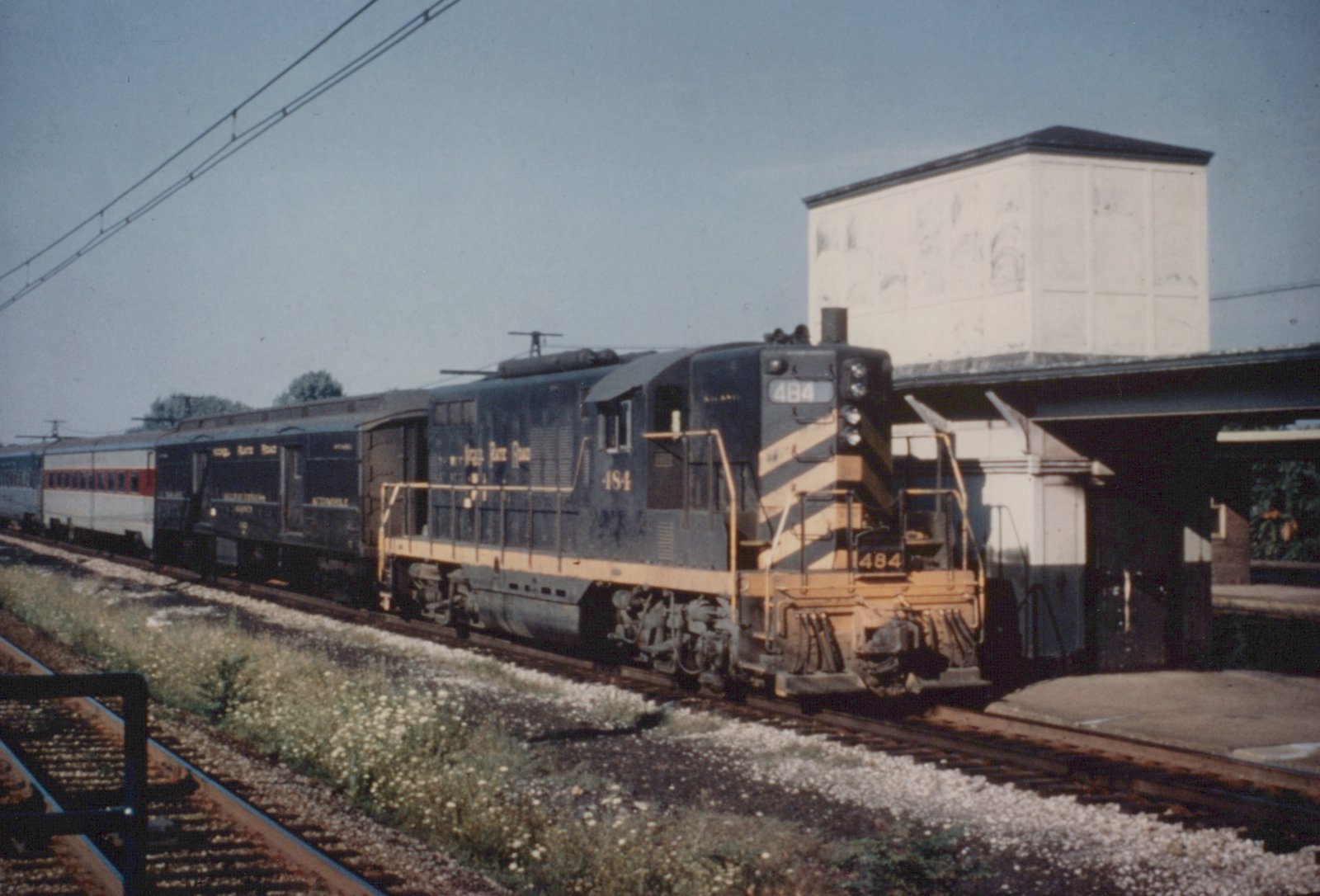 A blue and yellow New York Central GP9 diesel locomotive, numbered 484, is pulling a stream of silver passenger cars along a track next to a station platform under a clear sky.