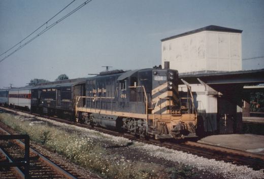 A blue and yellow New York Central GP9 diesel locomotive, numbered 484, is pulling a stream of silver passenger cars along a track next to a station platform under a clear sky.