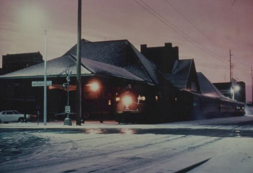 A black diesel locomotive with a bright headlight and the number 4019 on its front is stopped at a grade crossing on a snowy evening, with a station building and a street sign that reads 'SPRINGFIELD' in the background.