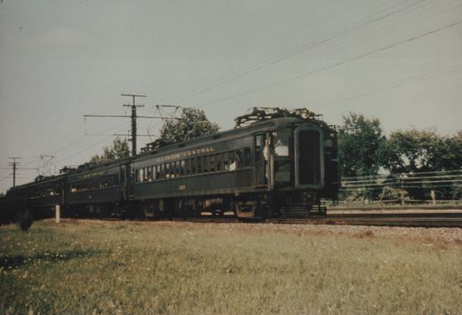 A dark green Illinois Central electric train with the number 1207 is moving on tracks beside a grassy field with overhead wires and trees in the background.