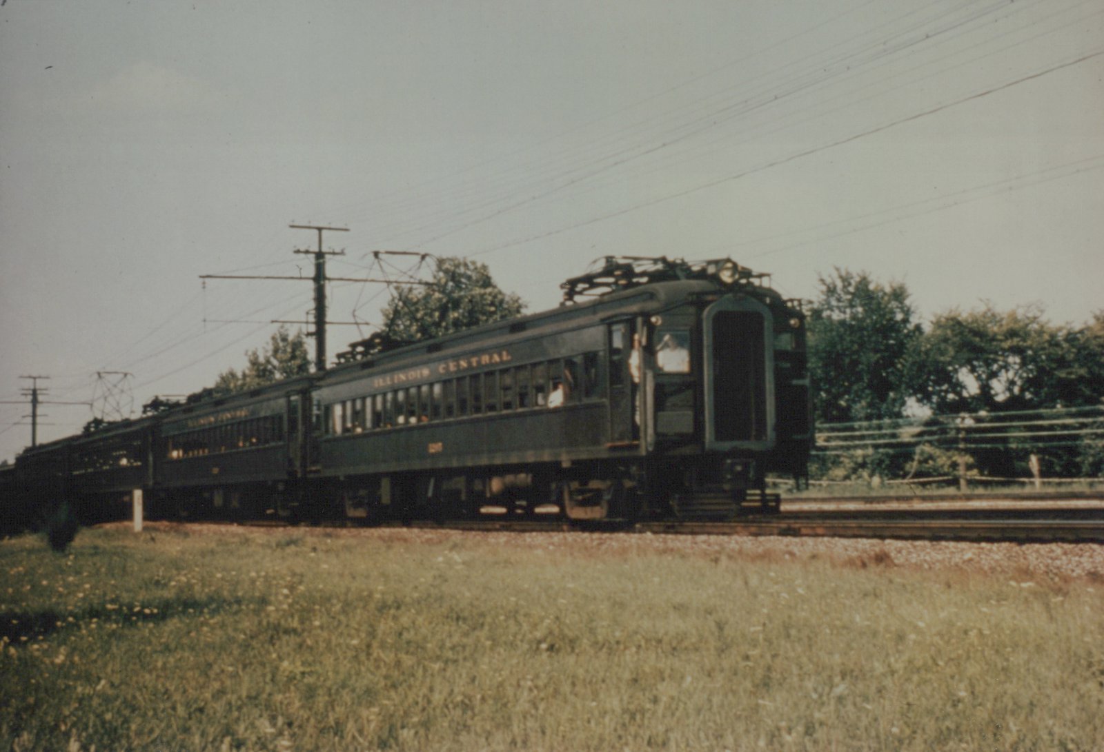 A dark green Illinois Central electric train with the number 1207 is moving on tracks beside a grassy field with overhead wires and trees in the background.