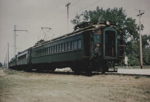 A vintage Illinois Central electric train with dark green, steel-bodied cars and pantographs, traveling along a track beside dry grass and utility poles on a summer day.