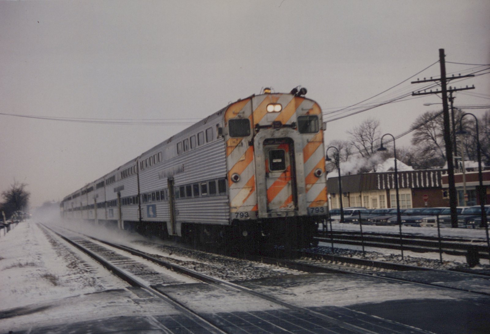 A silver Metra commuter train with an orange-and-white striped rear end is seen moving on snow-covered tracks, with bare trees and buildings in the background.