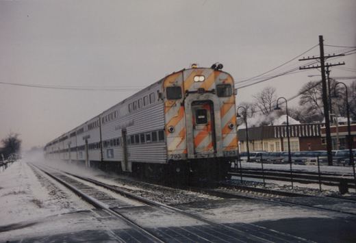 A silver Metra commuter train with an orange-and-white striped rear end is seen moving on snow-covered tracks, with bare trees and buildings in the background.