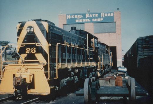 A black and yellow Nickel Plate Road diesel locomotive numbered 26 is parked on a track in front of a brick building with a sign reading 'Nickel Plate Road Diesel Shop.'