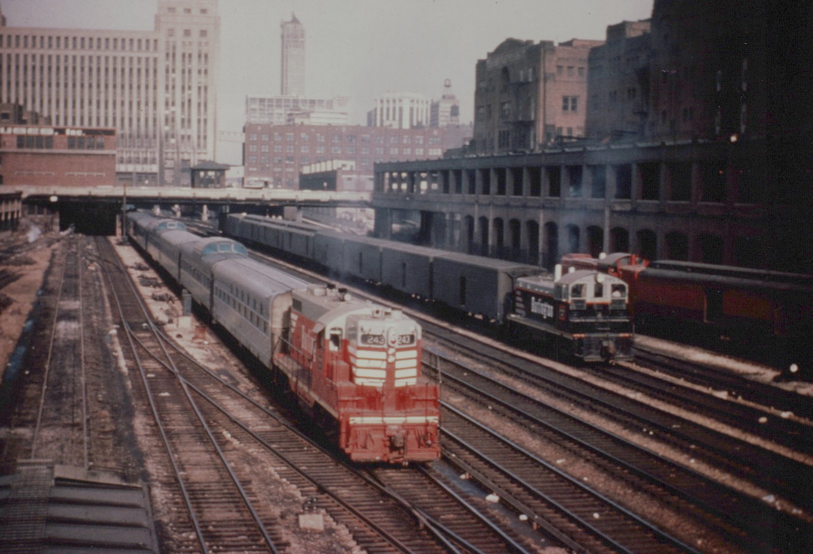 Two diesel locomotives and passenger cars on multiple tracks with city buildings and overpasses in the background.