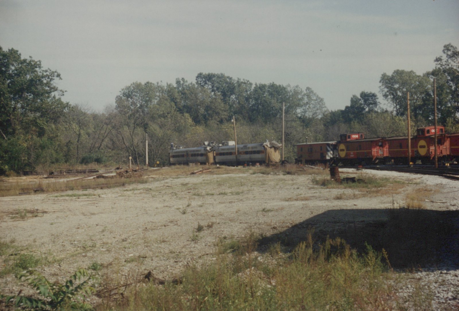 A photograph of a rail yard with several train cars parked on separate tracks. The ground is gravel and weeds, with trees in the background under an overcast sky.