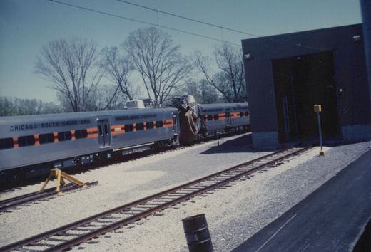 Two silver South Shore Line trains in a yard, one with a crumpled front end, beside a large dark building under a clear sky.