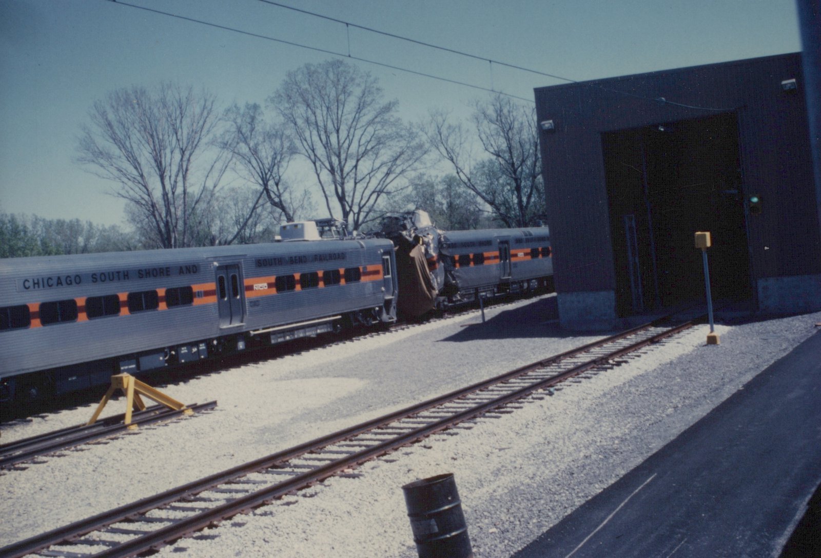 Two silver South Shore Line trains in a yard, one with a crumpled front end, beside a large dark building under a clear sky.