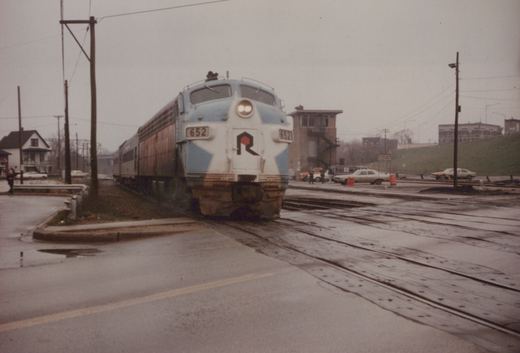 A blue and white diesel locomotive with the Rock Island 'R' logo on the front is crossing a wet street, with several cars parked nearby and a signal tower in the background.