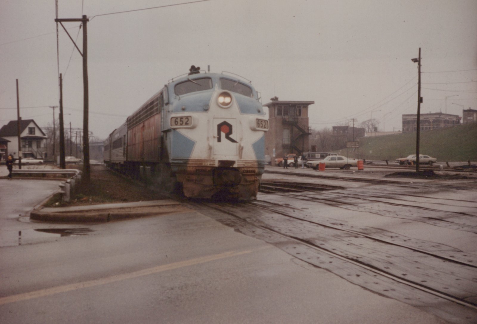 A blue and white diesel locomotive with the Rock Island 'R' logo on the front is crossing a wet street, with several cars parked nearby and a signal tower in the background.