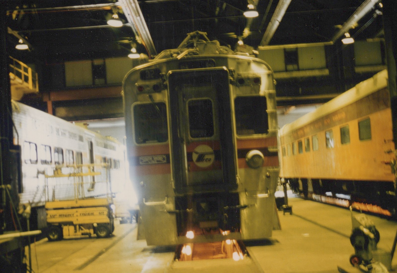 A front view of a silver and red South Shore Line train car, number 32, with an RTA logo, parked inside a well-lit train shop with other cars and equipment nearby.