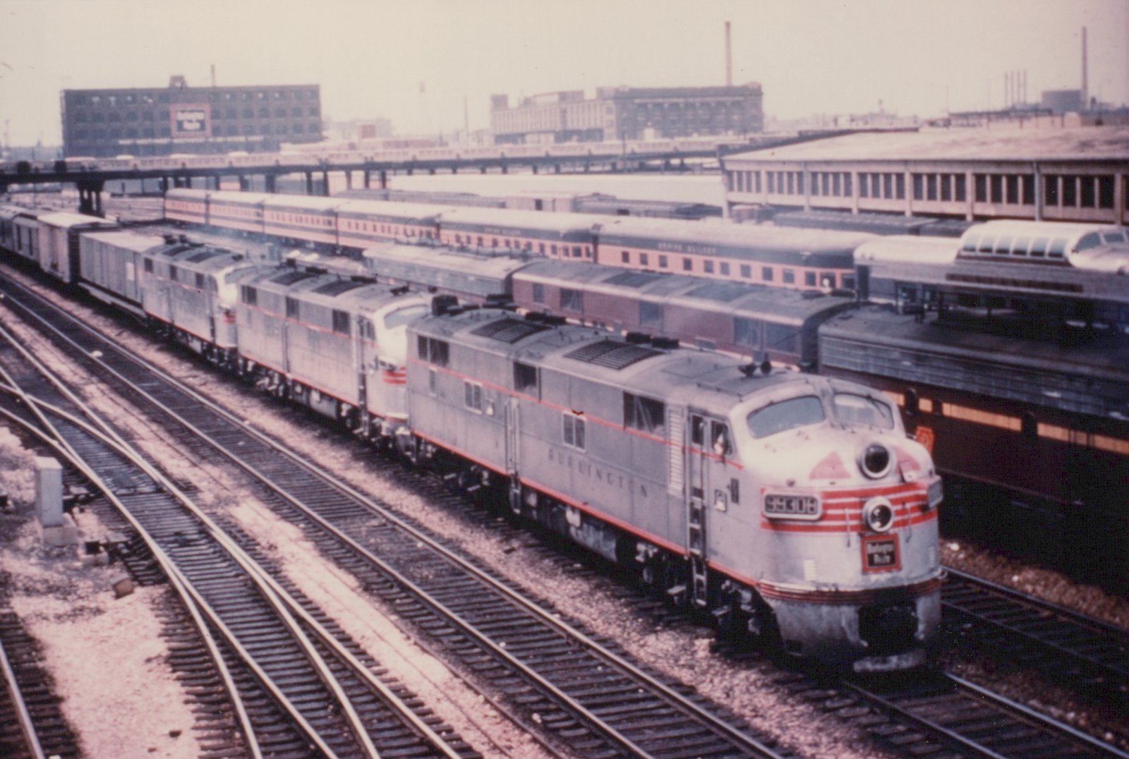 A silver EMD F3 diesel locomotive with red stripes, bearing the number 99306 and the name 'Denver Zephyr,' leads a train through a snowy yard with other passenger cars on adjacent tracks.