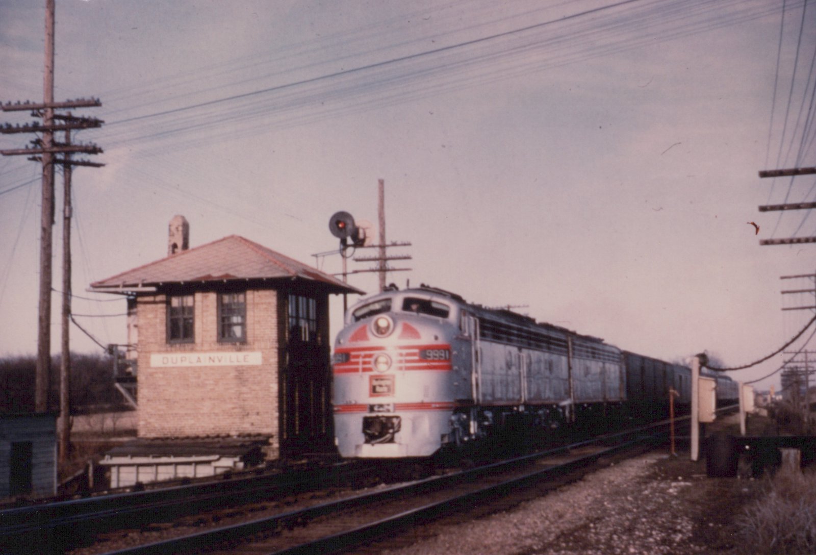 A silver and red Chicago Burlington and Quincy F7 diesel locomotive numbered 9991 pulls a line of freight cars past a brick signal tower with a sign reading 'DUPLA NVILLE' under an overcast sky.