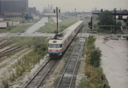 An elevated view of an Amtrak Metroliner train with a white, red, and blue livery moving on a wet, overgrown railroad track in an urban area.