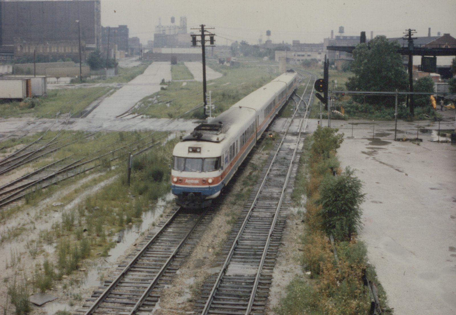 An elevated view of an Amtrak Metroliner train with a white, red, and blue livery moving on a wet, overgrown railroad track in an urban area.