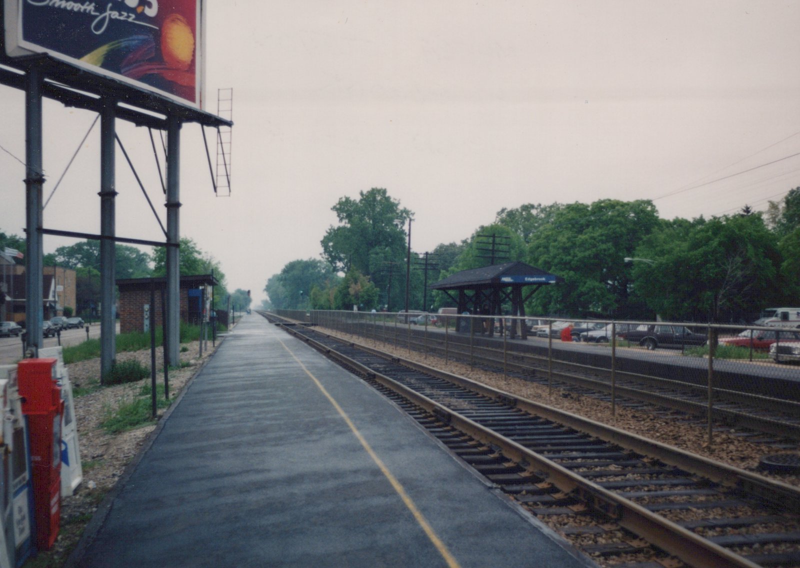 An empty train station platform with a large billboard for 'Smooth Jazz' on the left and a shelter with a blue 'Edgebrook' sign on the right, under an overcast sky.