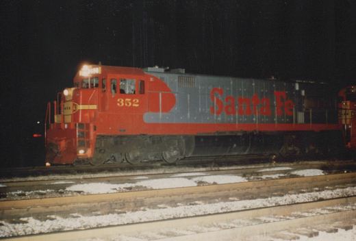 A red and silver Santa Fe diesel locomotive, number 352, sits on snowy tracks at night, its headlights on and the 'Santa Fe' name visible on its side.