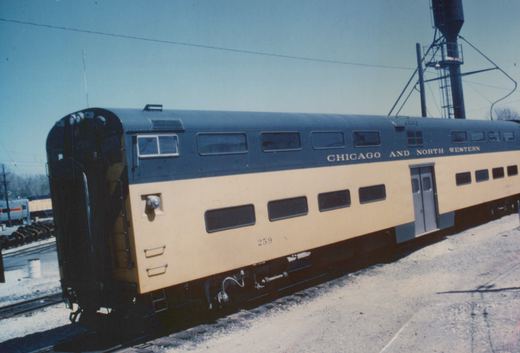 A side view of a two-level Chicago and North Western gallery passenger car, number 259, parked on tracks in a snowy rail yard under a clear blue sky.
