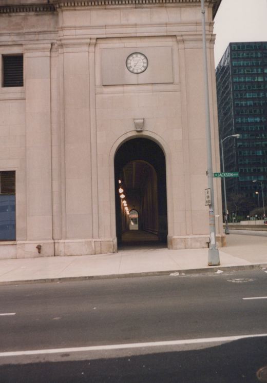 A large stone building with a clock above an arched tunnel entrance, with a street sign for W Jackson Blvd on a pole, and a modern high-rise building in the background.